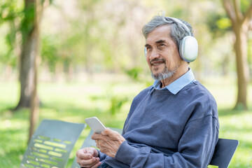 Fototapeta premium portrait happy asian senior man with grey hair and beard wears headset holding smartphone to listening music,sitting on bench in the park