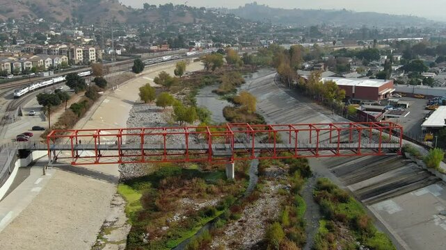 Aerial drone view of metal pedestrian bridge crossing the Los Angeles River showing concrete river channel surrounding city infrastructure industrial areas residential zones and distant hills