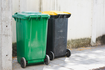 two large wheeled garbage bins placed on a sidewalk next to a wall. One bin is green with a green...