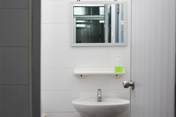 The bathroom has clean white tiles, a sink with a mirror, and a small shelf that looks neat and tidy.