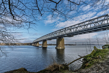 Obraz premium Steel Truss Bridge Spanning Wide River with Blue Sky and Leafy Foreground, Little Belt, Denmark