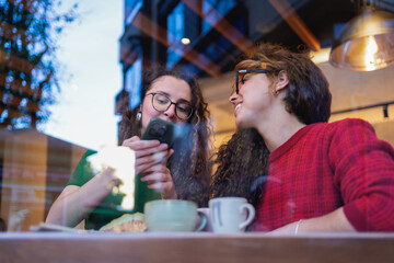 Two young women sharing a moment of connection over coffee and social media in a cozy cafe