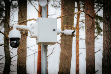 A close-up of a modern surveillance camera system mounted on a metal pole, featuring a dome camera and a bullet camera. The background includes trees and a security fence.
