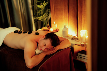 Hot stone massage at spa salon in luxury resort with warm candle light, blissful man customer enjoying spa basalt stone massage glide over body with soothing warmth. Quiescent