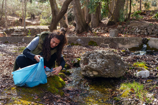 Young woman collecting plastic trash in blue bag from forest stream, environmental pollution problem