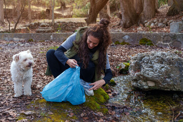 Volunteer collecting plastic bottle from a stream in a forest with her dog, environmental awareness concept
