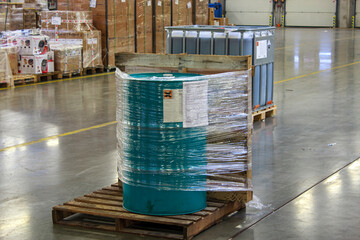 Green barrel wrapped in plastic and secured to a wooden pallet. Hazardous goods storage.Barrel is surrounded by packaged goods and another large container, indicating an organized storage environment.