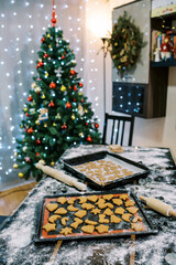 Baking trays with raw and baked cookies stand on a table next to a decorated Christmas tree