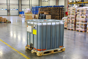 Hazardous goods storage. A large container filled with liquid is placed on a wooden pallet in a spacious warehouse. Shelves and stacks of packaged goods are visible in the background.