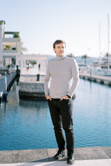 Fototapeta premium Young man with his hands in his pants pockets stands on the embankment near the moored yachts