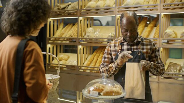 Senior Black baker in apron and gloves, wrapping fresh croissants in paper bag and selling pastries to female customer in bakery shop