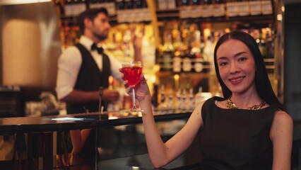 Beautiful guest looking at camera drinking at luxurious counter bar in mixed vodka Cosmopolitan red cocktail decorated with flower on bartender background in perfect atmosphere at nightclub. Vinosity.