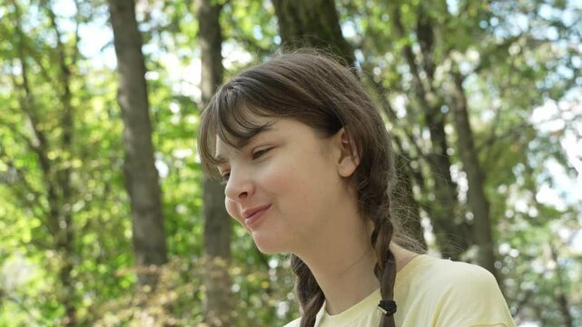 A teenage girl with pigtails in a yellow t-shirt smiles sweetly and wipes her hands with a wet napkin after lunch in a city park on a sunny summer day. Left-side shot, close-up