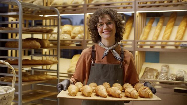 Happy female baker in apron and gloves holding wooden tray filled with freshly baked croissants, smiling at camera in cozy bakery with shelves of bread in background. Video portrait
