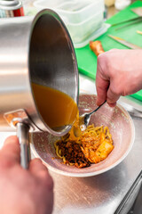 A chef pours rich broth over a bowl filled with noodles, vegetables, and meat, showcasing a culinary preparation. The vibrant colors and inviting presentation enhance the dish's appeal.