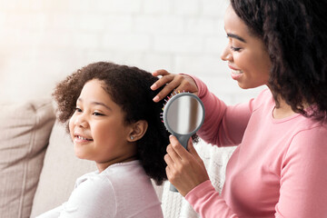 Pregnant afro woman brushing her cute little daughter's hair, sitting on couch at home, bonding...