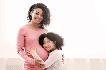 Happy expecting mom and her little afro daughter hugging next to window at home, looking at camera, copy space