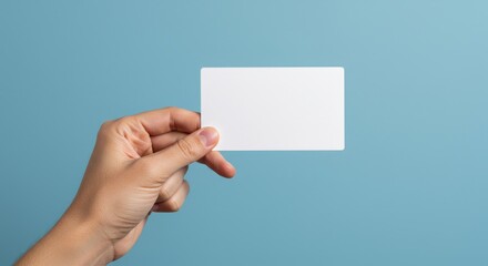 Fototapeta premium Hand Holding Blank White Card - Close-up of a hand holding a blank white card against a light blue background. Perfect for mockups and presentations