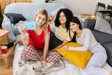Three cheerful young women taking a selfie with a smartphone while enjoying a slumber party at home, wearing pajamas and having fun. Female friendship and youth concept