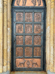 A bronze church door with engraved religious scenes, framed by a stone archway with intricate carvings. The door features biblical figures and symbolic imagery