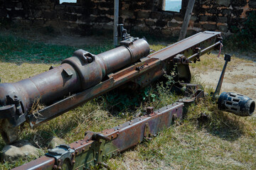 A decayed and rusted artillery cannon lies abandoned in an overgrown field near ancient stone ruins, representing remnants of historical warfare and military history.