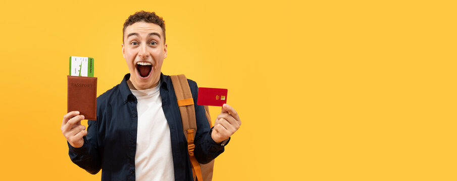 Happy excited guy student showing red plastic bank credit card and passport with flight tickets, carrying backpack, going vacation, travelling abroad, yellow studio background