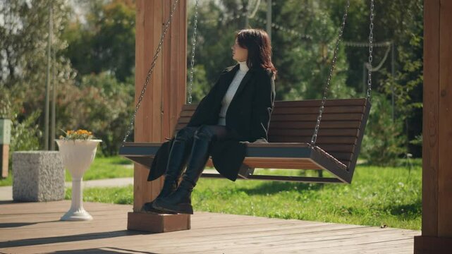 Lady in black coat and leather boots sitting on wooden swing chair in a public park, enjoying bright sunlight, with green park scenery and blurred background of trees and flowers