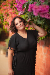 Plus size woman walks on a summer day along the street among the flowers blooming along and posing against the wall