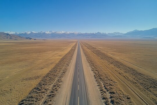 A long, straight highway cuts through a vast, arid desert landscape, with snow-capped mountains in the distance. - Powered by Adobe
