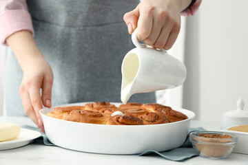 Woman adding frosting onto freshly baked cinnamon rolls at white table indoors, closeup
