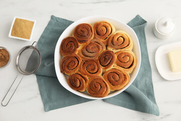 Tasty cinnamon rolls, ingredients and sieve on white marble table, flat lay