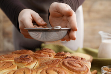 Woman with sieve adding cinnamon powder onto freshly baked rolls at table, closeup