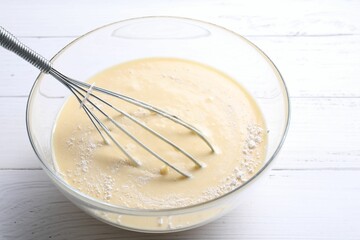 Whisk and bowl of dough on white wooden table, closeup