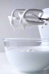 Hand mixer with whisks and bowl of whipped cream on white wooden table, closeup