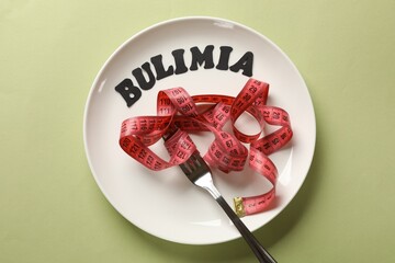 Plate with word Bulimia, measuring tape and fork on olive background, top view