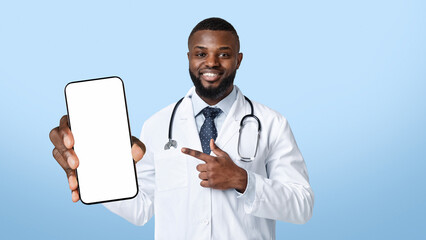 Smiling black physician with stethoscope pointing at mobile phone with blank screen, copy space