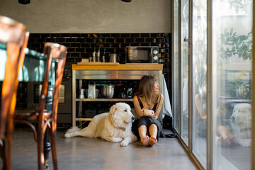 A barefoot woman sits on the floor of a modern kitchen, sipping coffee next to her large white dog. Natural light from glass windows enhances the peaceful and comforting atmosphere of the space
