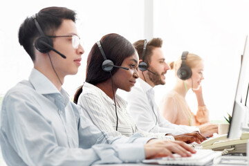 Line of call centre representatives with headsets working on computers at open space office. Group...