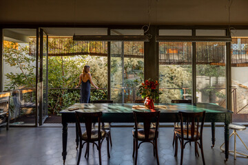 A woman in a sleek outfit stands on a contemporary balcony surrounded by lush greenery, enjoying the morning air. The open glass doors create a seamless connection between indoor comfort and outdoor