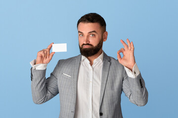 Confident corporate employee holding empty business card and showing ok gesture against blue background, space for design