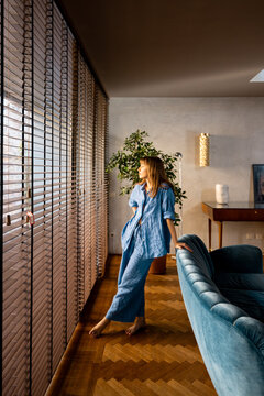 A woman in a relaxed blue linen outfit enjoys the warm morning light streaming through wooden blinds. The stylish interior, featuring a teal velvet sofa, wooden flooring, and elegant decor, conveys a
