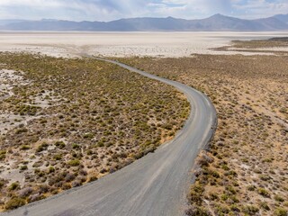 Fototapeta premium A winding gravel road cuts through the arid landscape of the Black Rock Desert in Nevada, USA. The road leads towards a vast, dry lakebed and distant mountains. Gerlach, Nevada, USA