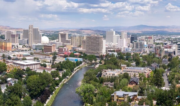 An aerial view of Reno, Nevada, USA, shows the Truckee River flowing through the city. The skyline features casinos and hotels, with mountains in the background.