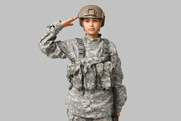Confident young woman in military uniform saluting on grey background