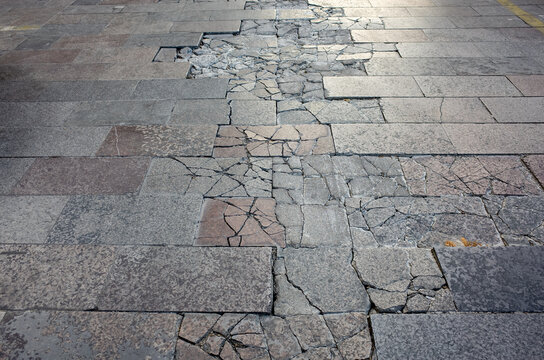 The fractured, uneven ground of a weathered pavement at an urban sidewalk or pedestrian walkway shows significant decay, with cracked and broken stone tiles scattered across the surface