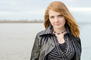 Young woman with red hair and light skin stands by calm lake. Wearing black leather jacket and...