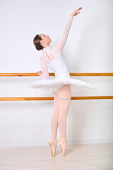 Fototapeta premium Young female ballet dancer with brown hair in white tutu poses gracefully on pointe. Bright studio with white walls and wooden bar emphasizes her form. Focus on elegance and poise