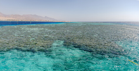 Panoramic view of Red Sea coral reef under bright daylight. Clear turquoise water reveals vibrant marine ecosystem. Mountains create distant backdrop, enhancing serene atmosphere