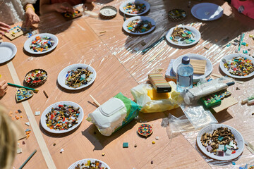 Sunlit workshop scene featuring multiple paper plates with vibrant mosaic tiles. Hands actively...