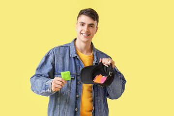 Young man holding cap with condoms on yellow background